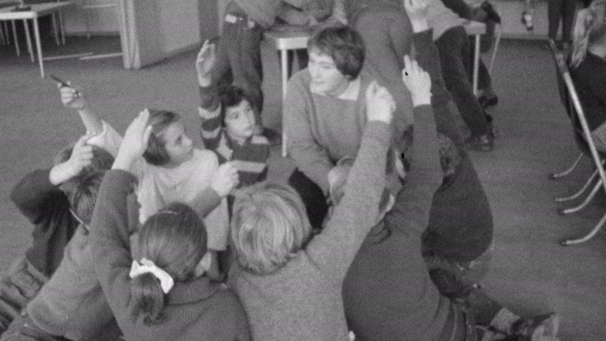 Black and white still of a teacher surrounded by young pupils seated with their hands up.