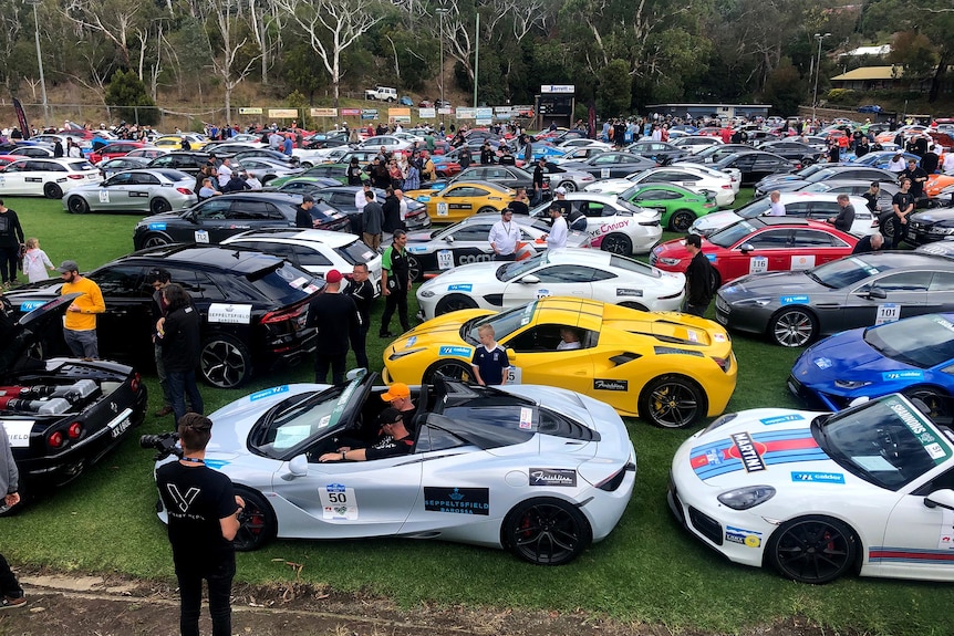 A bunch of cars park on an oval with people milling about.