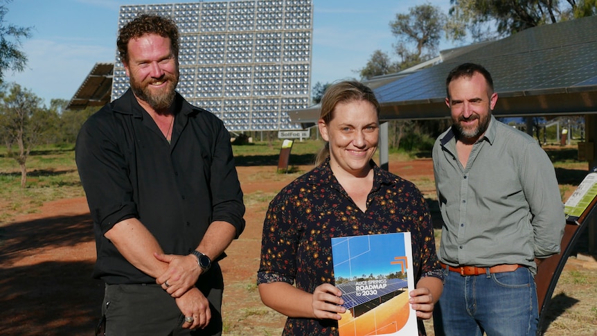 A woman holding the roadmap report stand in front of two men. They are in front of a solar panel.