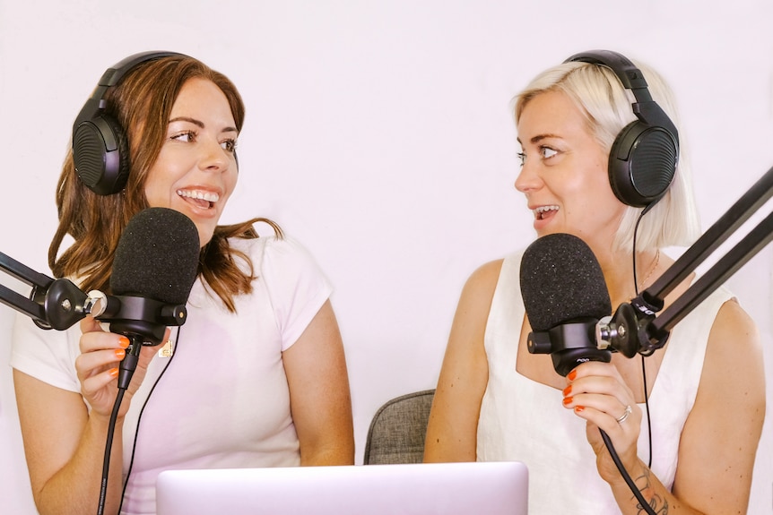 Two young women wearing headphones sit talking together at microphones.