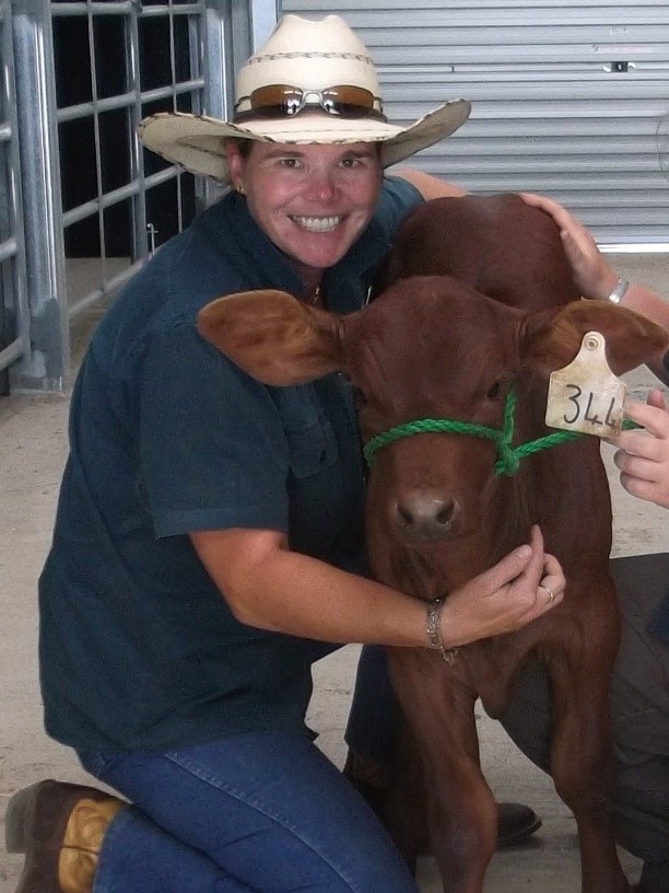 A woman wearing a white wide-brimmed hat kneeling on concrete smiles at the camera while patting a dark brown cow.