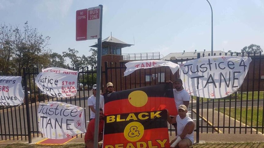 Five men stand in front of the prison gates surrounded by posters reading: Justice for Tane