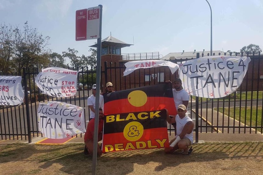 Five men stand in front of the prison gates surrounded by posters reading: Justice for Tane