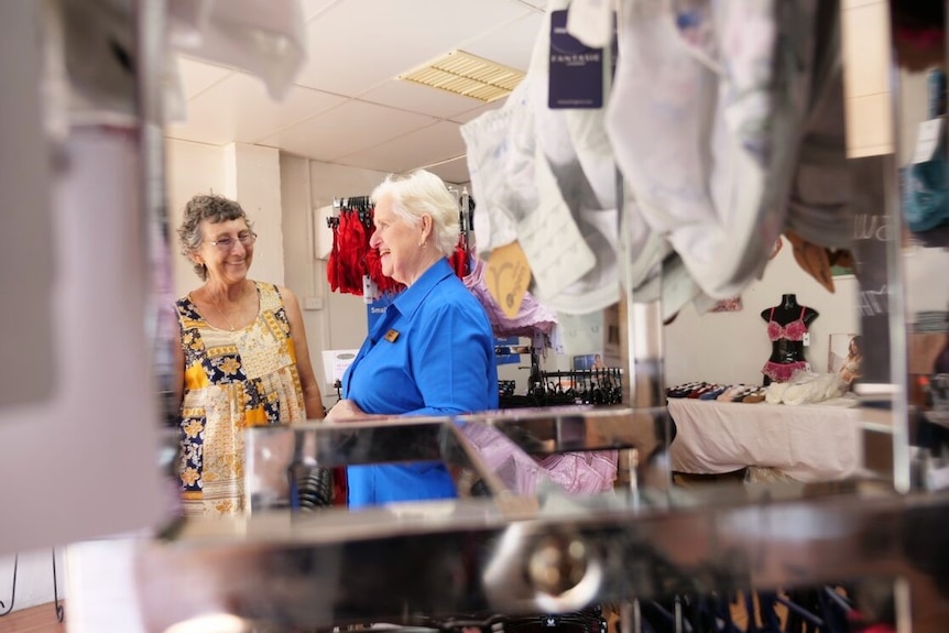 Two women seen chatting through a rack of bras. 