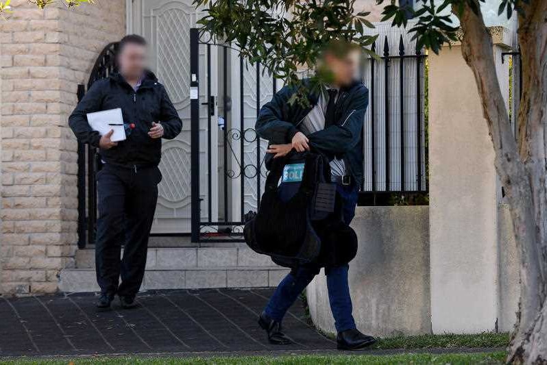 Two men wearing black and carrying bags walk away from the gate of a house.