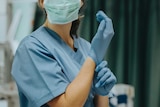 A masked nurse adjusts their blue plastic glove in an operation room.