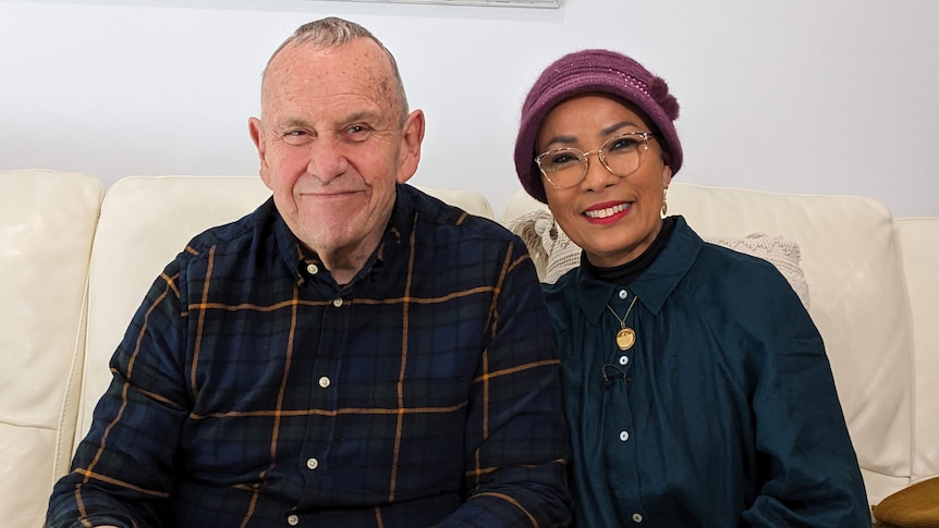 A grey haired man and woman sit smiling on a white couch.