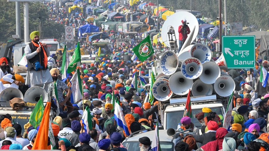 A wide shot shows a long road packed with vehicles and protesting farmers in colourful clothes.