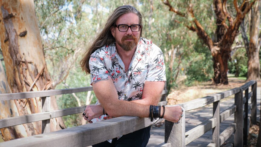 Nic Healey leaning on the rail of an old wooden bridge in a gum tree forest.