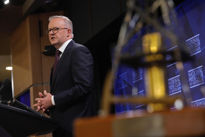 Anthony Albanese, a man in a dark suit and spectacles, stands at a lectern on a stage at the National Press Club