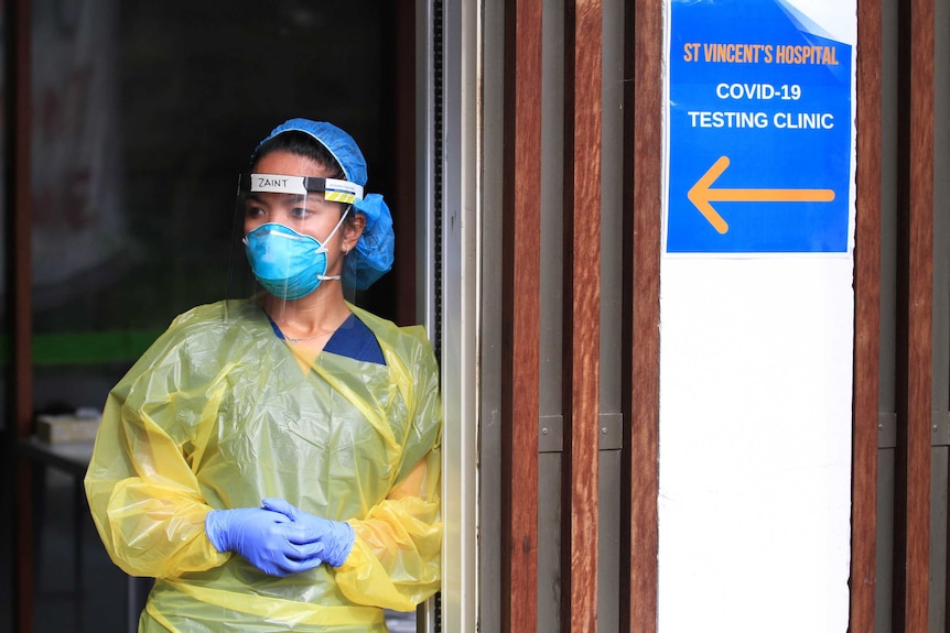 Woman wearing face mark and plastic visor with yellow plastic gown and blue gloves stands next to a COVID-19 testing clinic sign