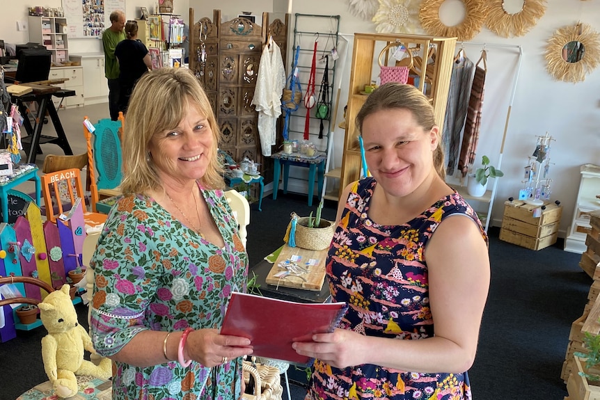 Two women in centre of shop scene holding folder, looking at camera, shop background has crafts and plants and furniture.