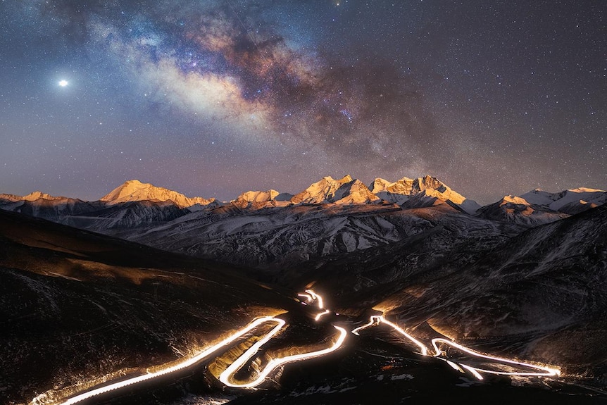 Vía Láctea sobre las montañas con una carretera iluminada al fondo. 
