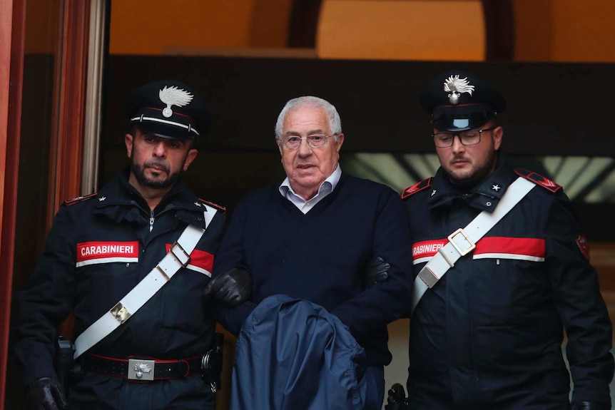 Two Italian police officers flank a handcuffed elderly man in glasses and a navy jumper.