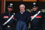 Two Italian police officers flank a handcuffed elderly man in glasses and a navy jumper.