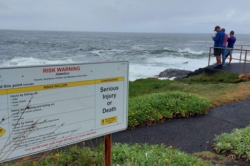 A warning sign near the ocean, with three people standing on the lookout on the right.