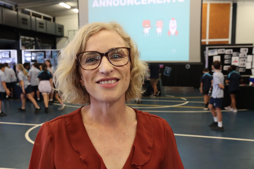 A blonde woman with glasses smiles at the camera. There is a group of school students behind her