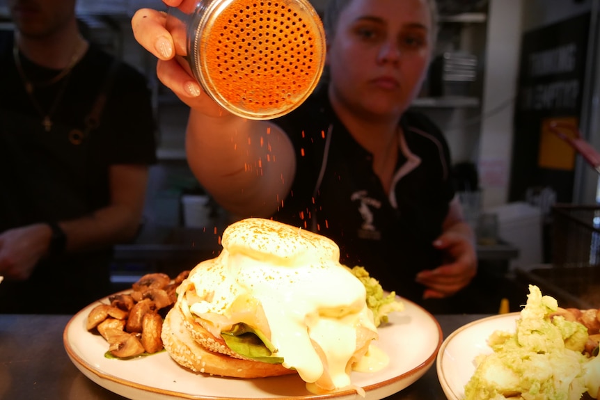 A young woman sprinkles paprika on a serving of avocado on toast with a poached egg
