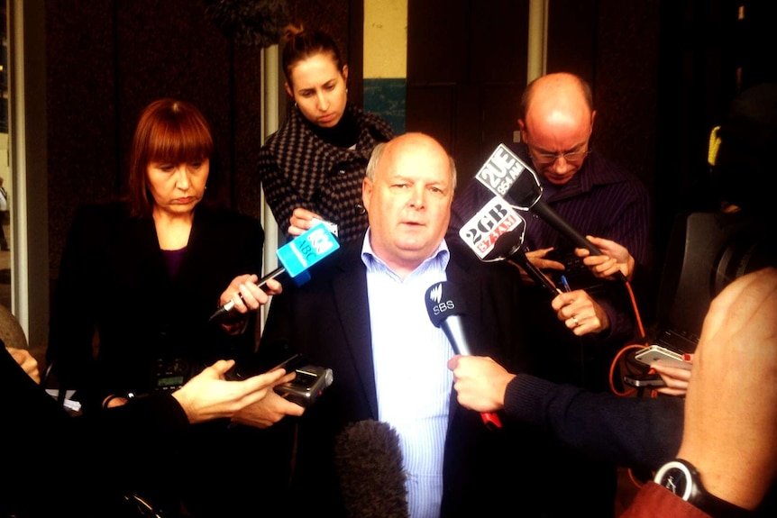 A media scrum outside court, reporters holding their microphones out towards a man in a suit.