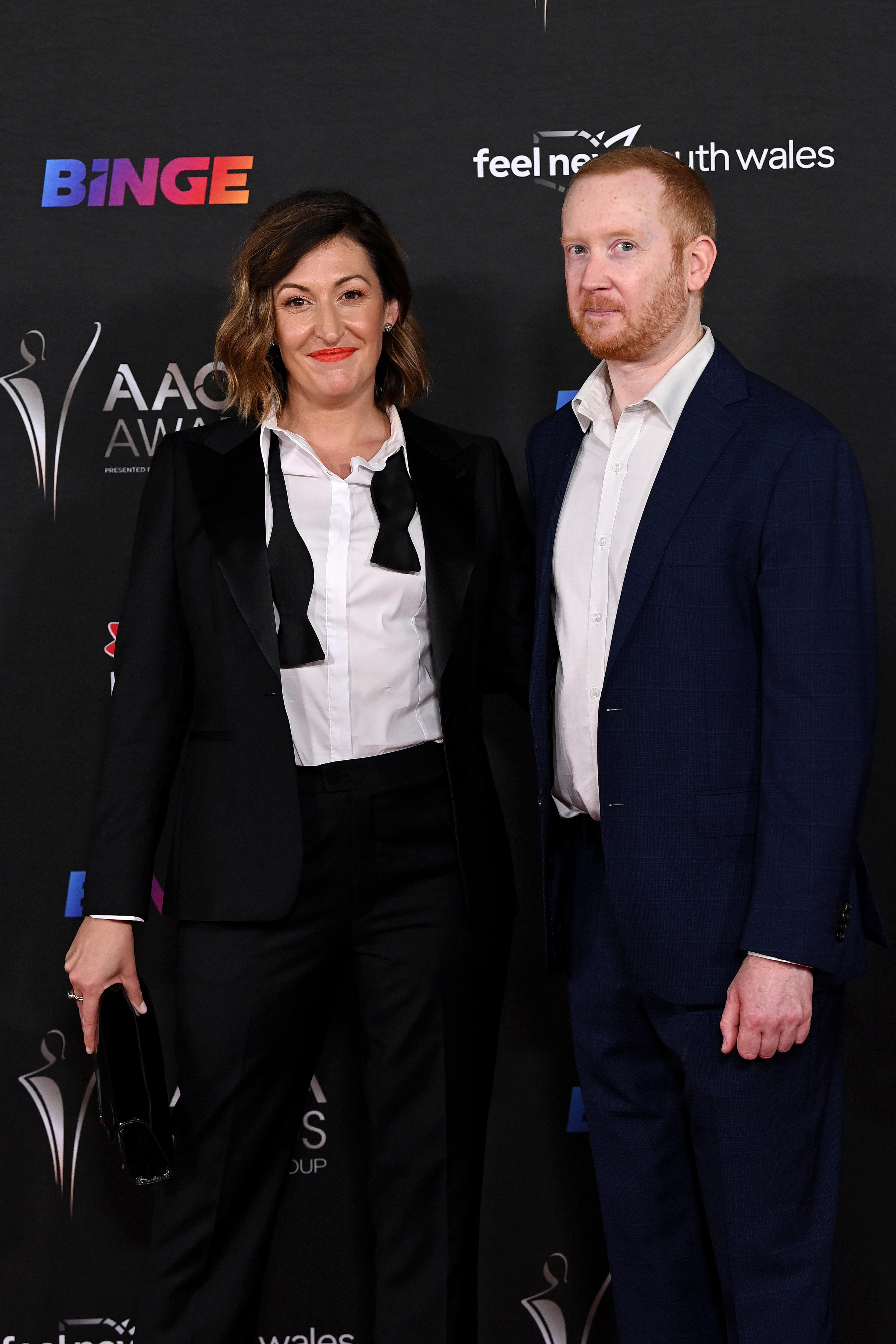 A woman with dark short hair in a black suit smiling beside a man with short orange hair in a suit, no tie