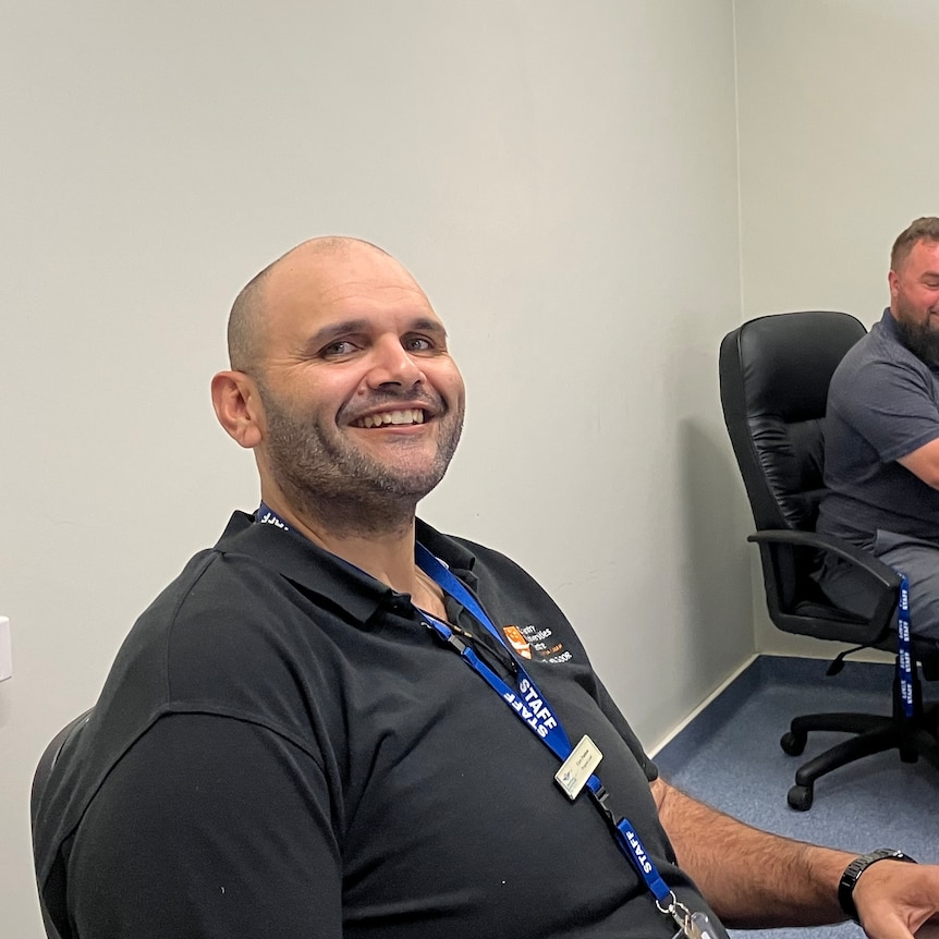 A bald indigenous man smiling while he sits in an office chair next one of his colleagues at the Royal Flying Doctors Service