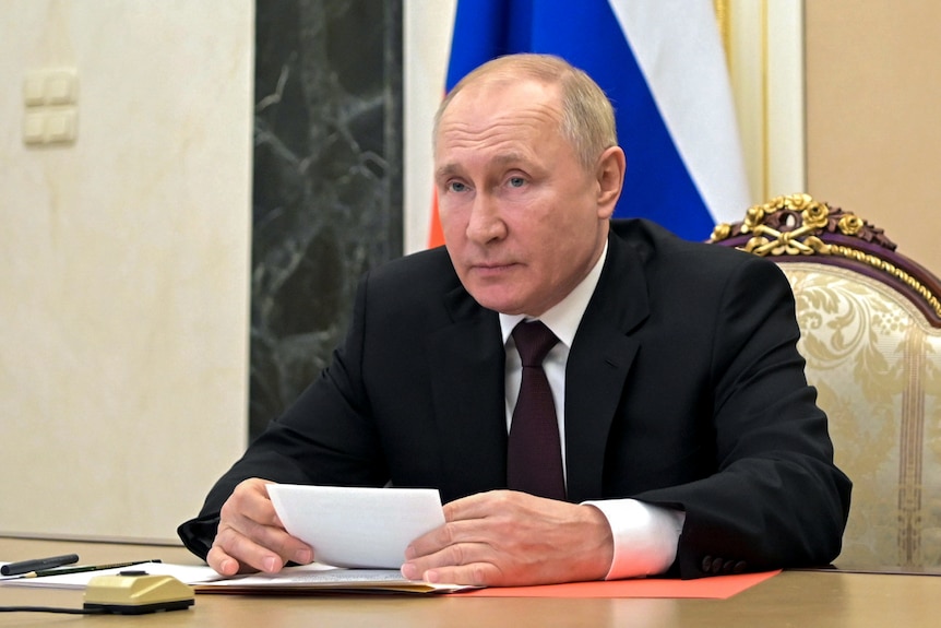 Vladimir Putin in a black suit and tie sitting at a desk and holding a piece of paper during a meeting in Moscow, Russia