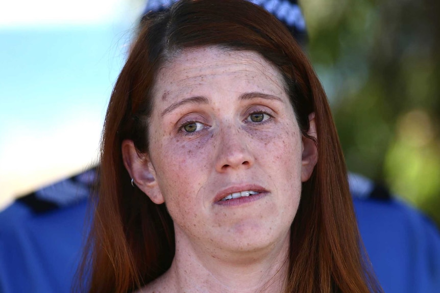 A young woman in a black singlet top looks emotional as police stand behind her.