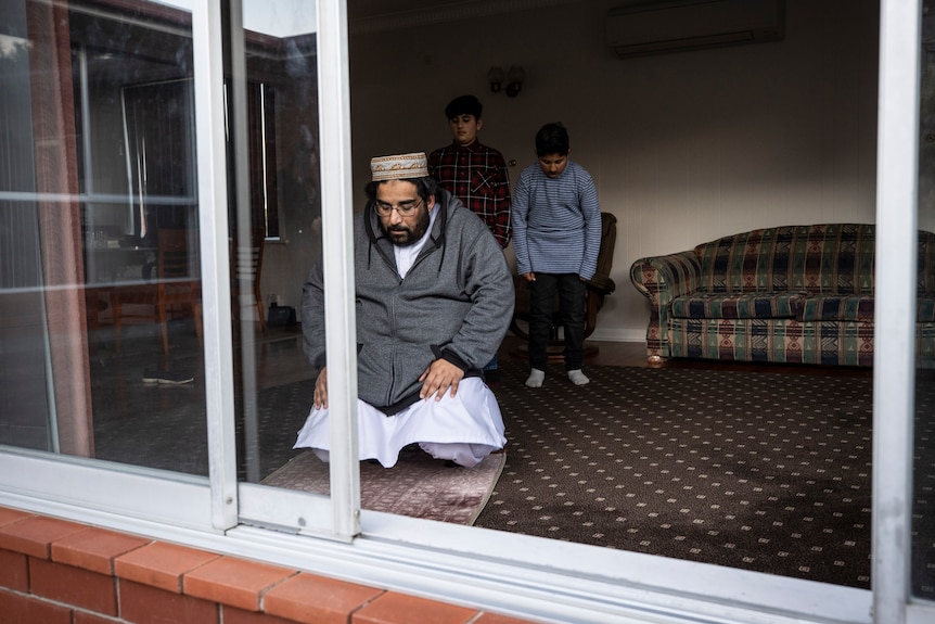 A father kneels on the living room floor and prays as his two sons stand behind him heads bowed. 