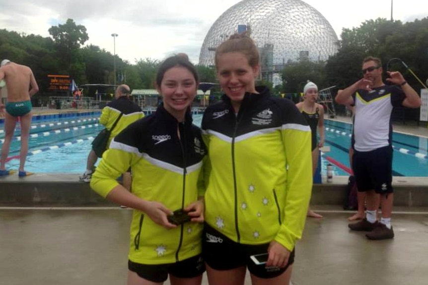 Two young women in matching black and yellow team uniforms stand and smile. Behind them is a swimming pool with lane 