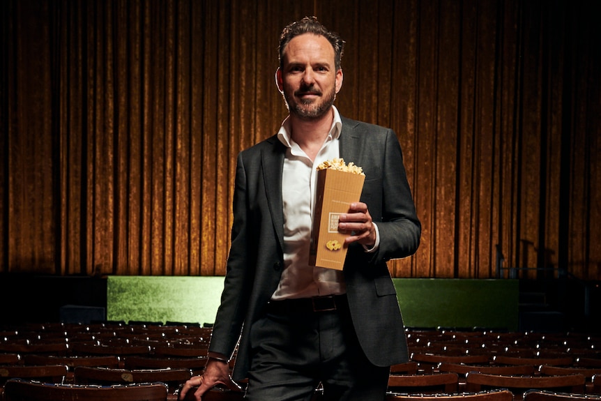 Benjamin Zeccola holding a carton of popcorn in a movie theatre, curtains behind him