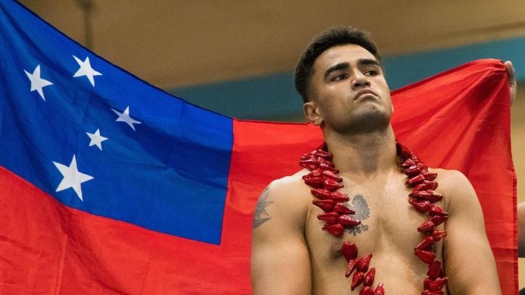Boxer Ato Plodzicki-Faoagali stands shirtless with blue boxing gloves on, infront of red and blue Samoan flag.