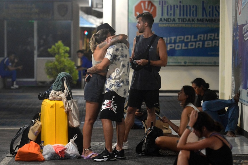 A group of tourists stand near luggage, with a woman and a man hugging as others sit nearby looking at mobile devices.