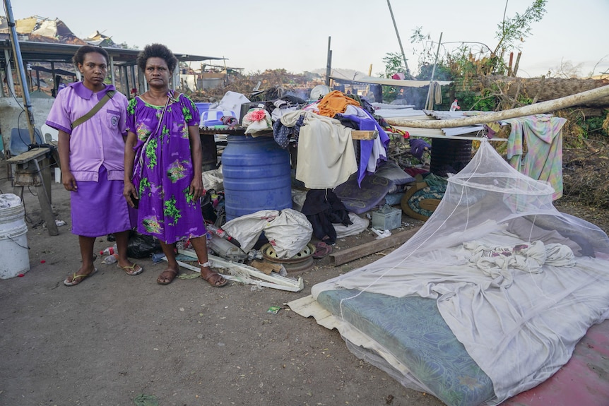 Two women stand next a mattress on the ground and a collection of belongings.