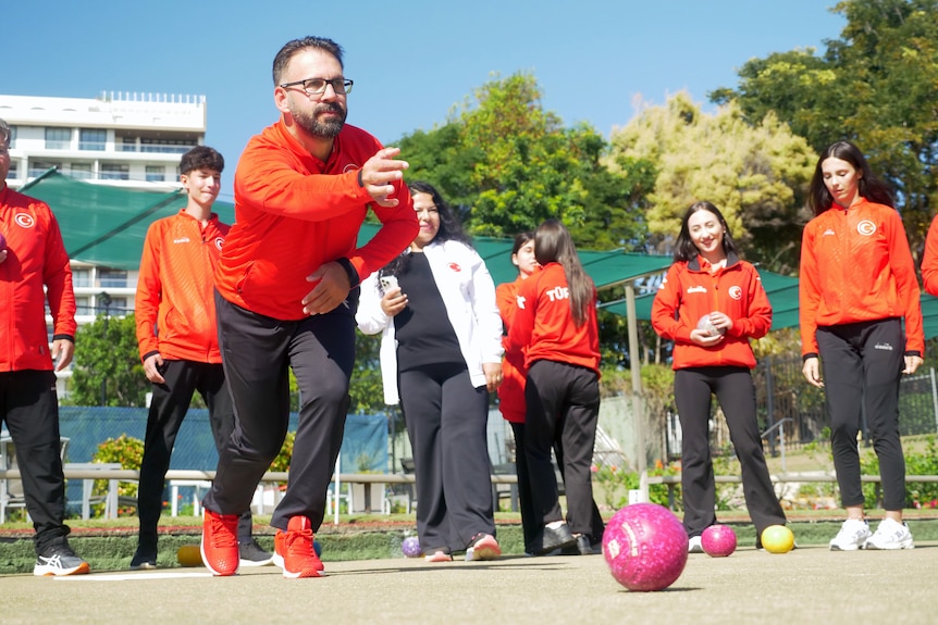 A man bowling a pink ball with people behind him, all dressed in orange top and black pants, blue skies, green trees.