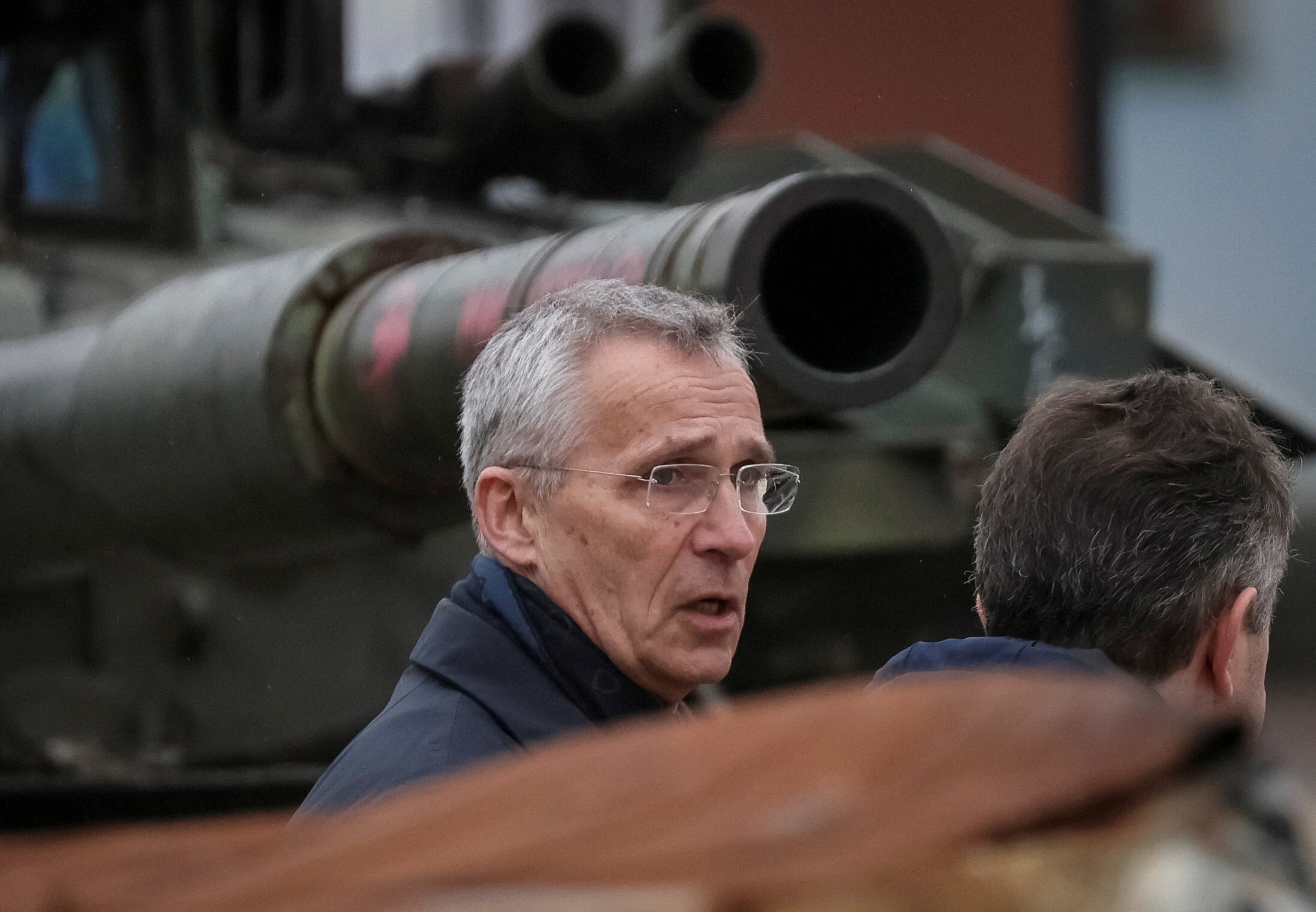 Jens Stoltenberg, a man with grey short hair and glasses, is captured speaking to the back on someone's head in front of a tank.