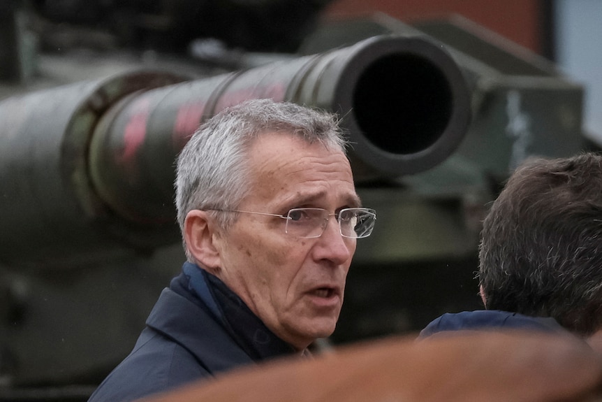 Jens Stoltenberg, a man with grey short hair and glasses, is captured speaking to the back on someone's head in front of a tank.