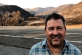 A man stands next to a road with charred landscape behind him