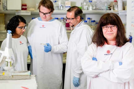 Gilda Tachedjian stands, arms folded in a white lab coat, in a lab in front of two men and one woman in lab coats.