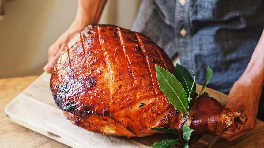 A ham leg glazed with maple syrup and spices and baked til its golden is on a wooden board being placed down leaves as garnish