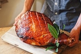 A ham leg glazed with maple syrup and spices and baked til its golden is on a wooden board being placed down leaves as garnish