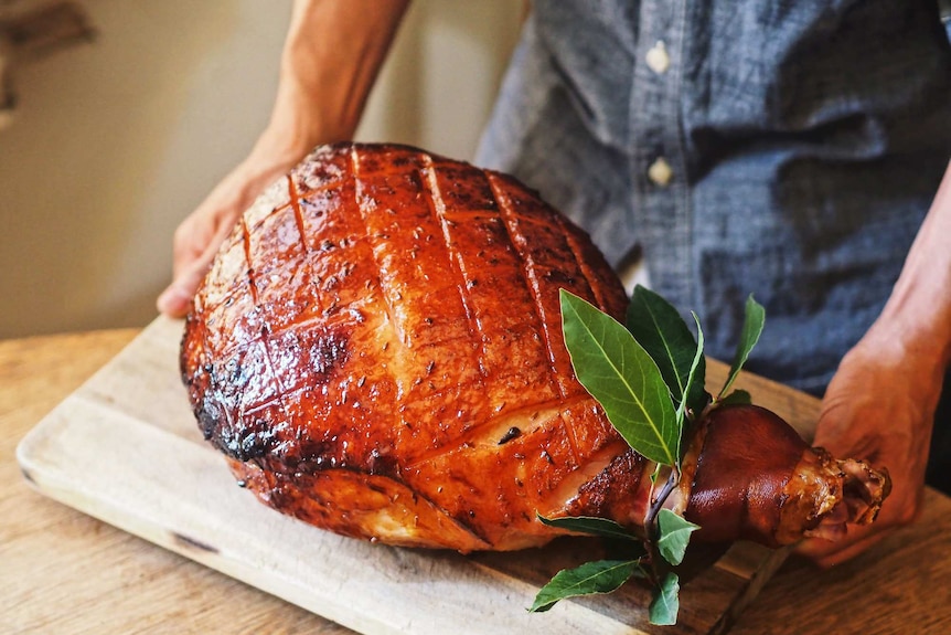 A ham leg glazed with maple syrup and spices and baked til its golden is on a wooden board being placed down leaves as garnish