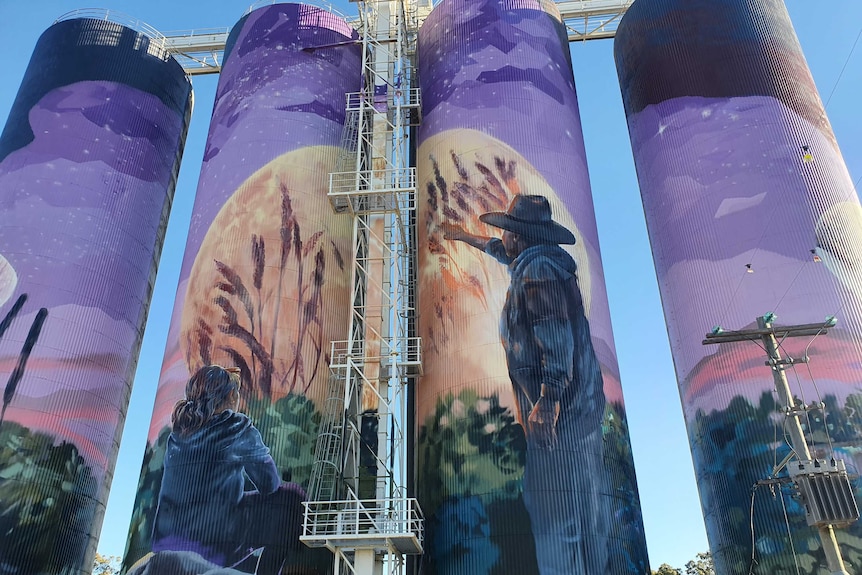 Four silos painted with a huge purple mural featuring a moon and a man in an Akubra-style hat.