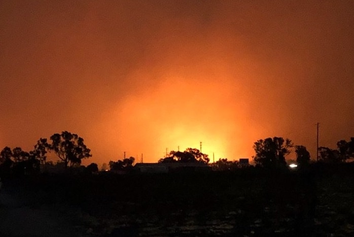 The glow from a bushfire at night, casting a silhouette against the horizon of trees and buildings.