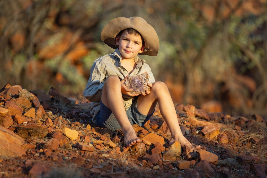 A 10 year old boy, sits on the red dirt and rocks barefoot holding a large rock with crystals.
