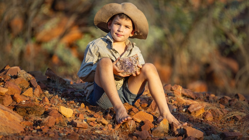 A 10 year old boy, sits on the red dirt and rocks barefoot holding a large rock with crystals.