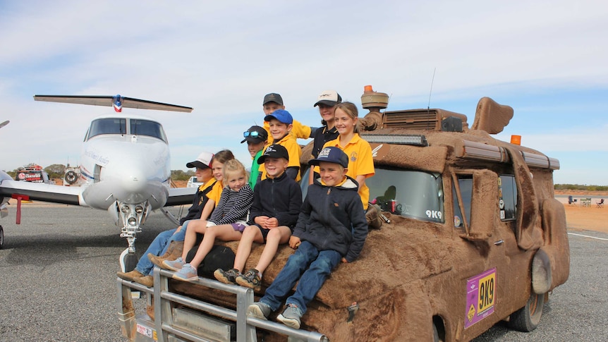 Students from Broken Hill School of the Airl sitting on the bonnet of a fur-covered van next to a RFDS plane.
