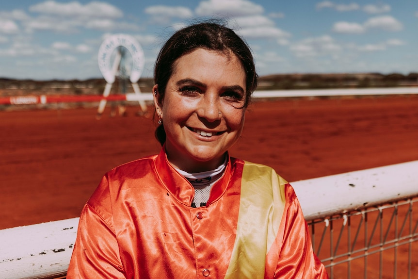 A female jockey posing in her racing colours near the finish line.