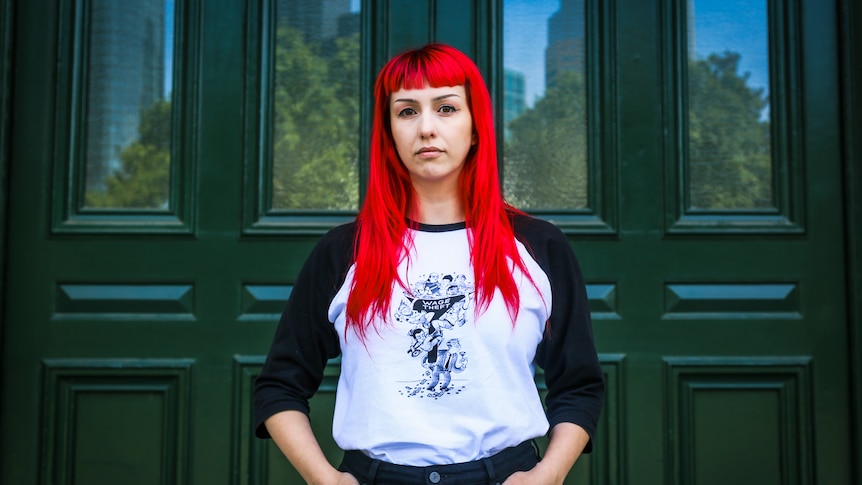 A woman with bright red hair has her hands in her pockets, standing in front of large green doors.