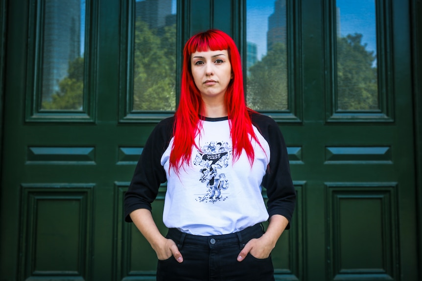 A woman with bright red hair has her hands in her pockets, standing in front of large green doors.