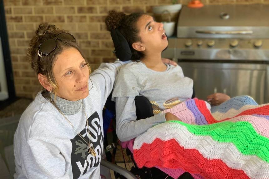 Lacey Harrison sits with her arm around her daughter Denishar Woods, who is in a mobile chair with a blanket on her lap.